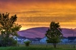 Stock photo of a fiery orange and gold sunset with cloud formations over the City of Winnipeg, Manitoba, Canada. The hills appear purple behind the foreground of grass and large trees along a fence with the white buildings of the city in the distance at t