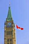 Stock photo of the top portion of the Peace Tower photographed showing the tall flagpole with Canadian National Flag right beside the tower. A background of clear light blue sky highlights the detail of the Peace Tower's structure including its windows, c