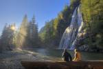 Stock photo of a man and a woman sitting on a log enjoying the scenic view of Virgin Falls near Tofino on Vancouver Island with sun rays filtering through the rain forest.