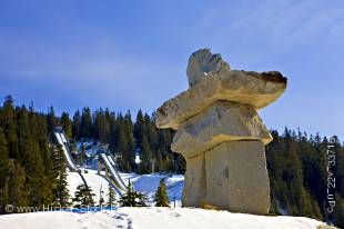 Stock photo of a Large stone inukshuk - Ilanaaq (the Vancouver 2010 Olympic Winter Games emblem) at the entrance to the Whistler Olympic Park Nordic Sports Venue, with the Olympic Ski Jumps and blue sky in the background, Callaghan Valley, British Columbi