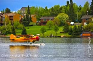 Stock photo of a bright orange Norseman aircraft taxiing on the water in the town of Red Lake, Ontario, Canada. The shore of the lake across the water has many beautiful trees and some brick buildings under a bright blue sky.