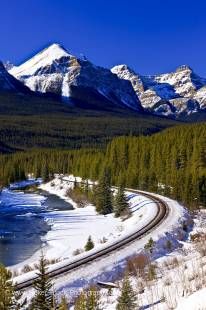 Stock photo of railway tracks beside the snow and ice fringed Bow River during winter with Fairview Mountain and blue sky in the background, Banff National Park, Canadian Rocky Mountains, Alberta, Canada. 